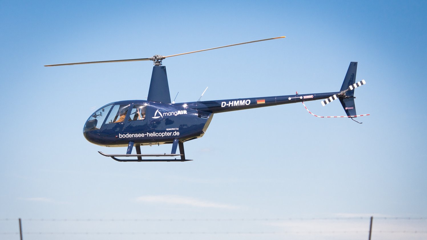 Dunkelblauer Hubschrauber mit weißem Schriftzug fliegt über einer Wiesenlandschaft unter blauem Himmel.
