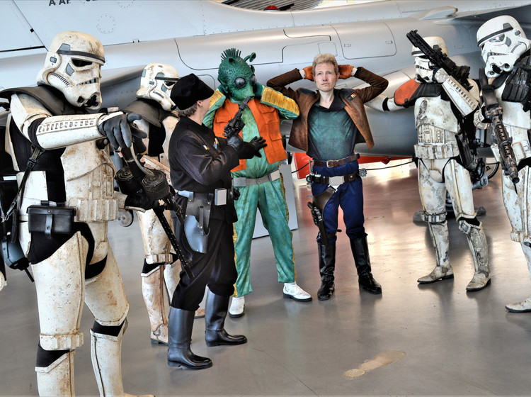 Mehrere Personen in Star-Wars-Kostümen im Hangar mit Flugzeugen im Hintergrund.