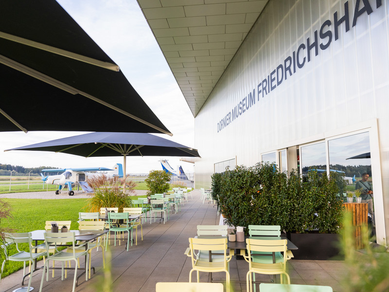 Außenterrasse mit bunten Tischen und Stühlen unter großen Sonnenschirmen vor dem Gebäude des Dornier Museums, im Hintergrund ein Flugzeug und grüne Wiesen.