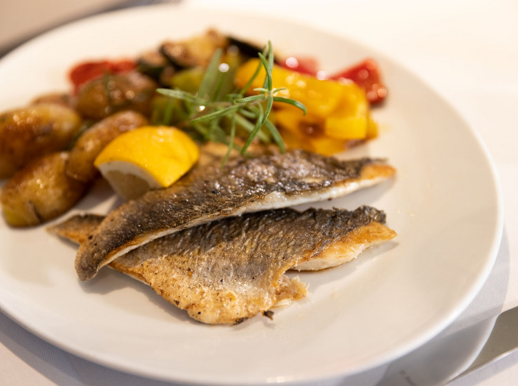 Servierter Teller mit gebratenem Fischfilet, Rosmarinkartoffeln, Zitronenspalte und buntem Ofengemüse auf weißem Teller.