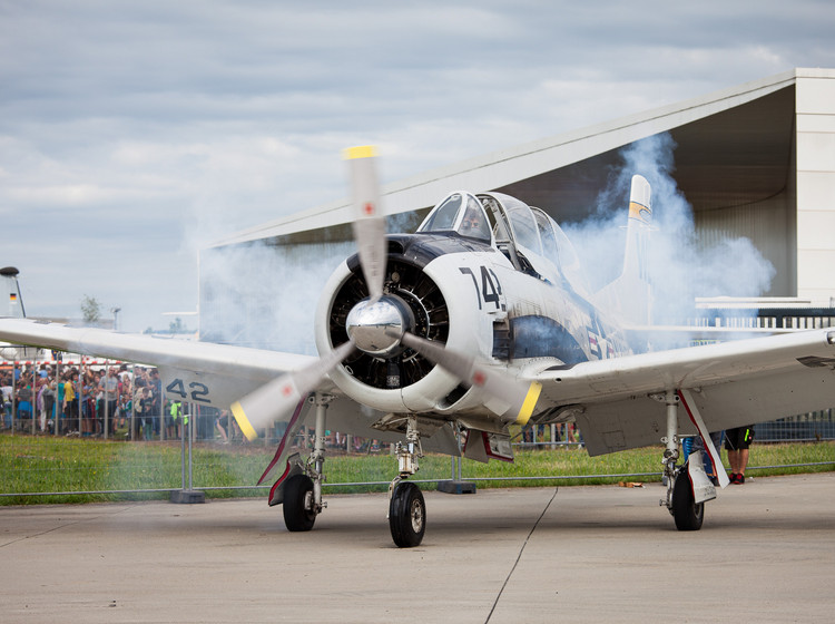 Ein Propellerflugzeug beschleunigt auf der Startbahn, dabei kommen Rauchschwaden aus den Triebwerken.