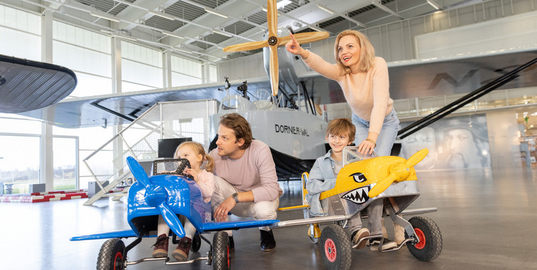 A Family of four in the Hangar of the Dornier Museum with a plane in the background. The two children are sitting in little toy planes.