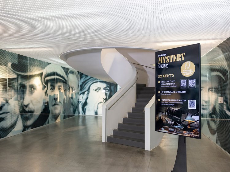 Infotafel zur Dornier Mystery Tour im Foyer des Dornier Museums mit Wendeltreppe und historischen Wandbildern.
