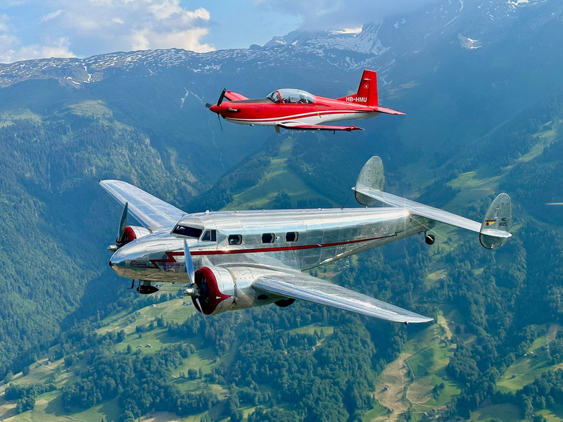 Zwei kleine Flugzeuge fliegen nebeneinander am Himmel, aus einer seitlich erhöhten Perspektive betrachtet mit einer Berglandschaft im Hintergrund.