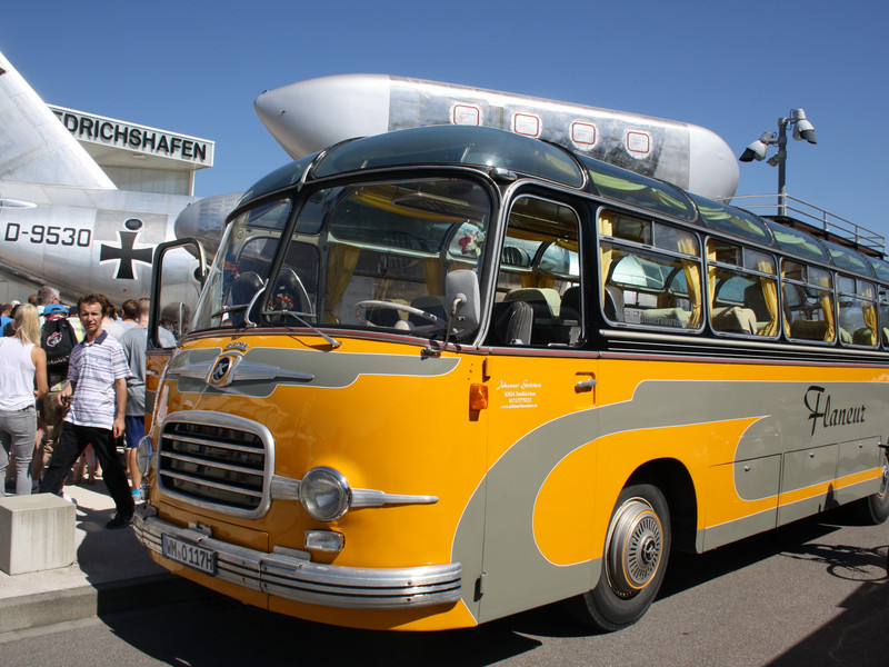 Gelber historischer Reisebus mit offenem Dach und Passagieren, der vor einem großen weißen Flugzeug auf einem Flugplatz steht.