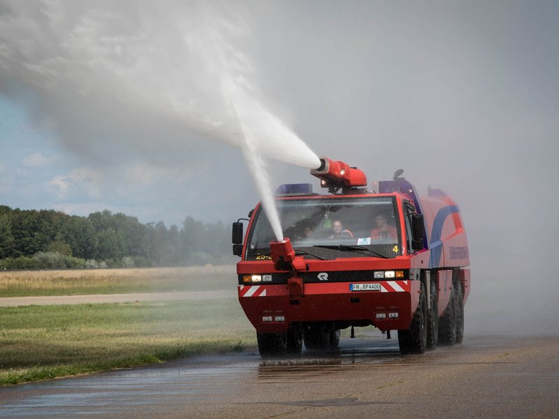Ein Flughafen-Feuerwehrfahrzeug von Vorne spritzt große Wassermassen auf das Rollfeld.