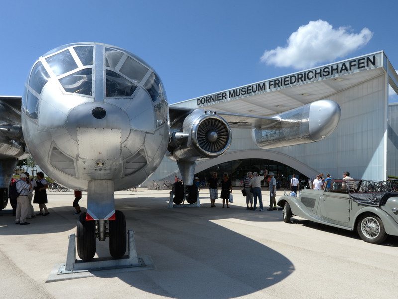 Historische Flugzeuge auf dem Außengelände des Dornier Museums bei Sonnenschein, darunter ein großes silbernes Passagierflugzeug mit Besuchern davor
