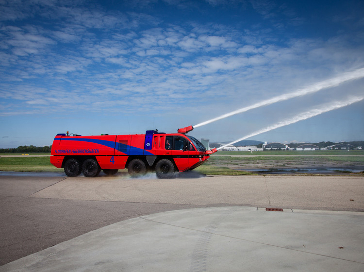 Rotes Feuerlöschfahrzeug auf Asphalt fährt auf einer Flugplatzpiste und sprüht Wasserstrahlen in den blauen Himmel.