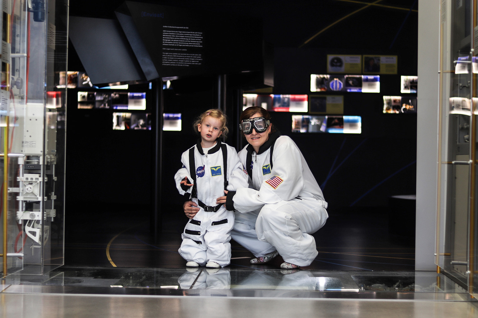 Zwei Kinder in weißen Astronautenanzügen stehen und knien vor einer dunklen Raumfahrtausstellung mit beleuchteten Bildtafeln.