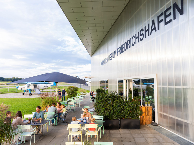 Mehrere Personen sitzen an den Tischen im Außenbereich des Dornier-Restaurants direkt am Rollfeld.