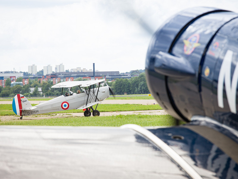 Ein Flugzeug mit sich drehendem Propeller fährt von rechts in das Bild, während im Hintergrund weiteres Flugzeug auf der Startbahn steht.