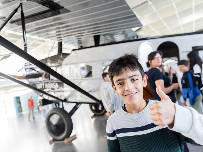 Ein junge hält den Daumen hoch, im Hintergrund steht ein Flugzeug im Hangar.