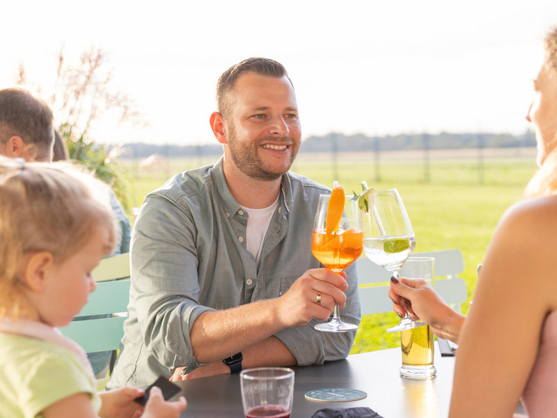 Ein Mann und eine Frau stoßen mit Cocktails an, während ein Kind links neben ihnen am Tisch sitzt.
