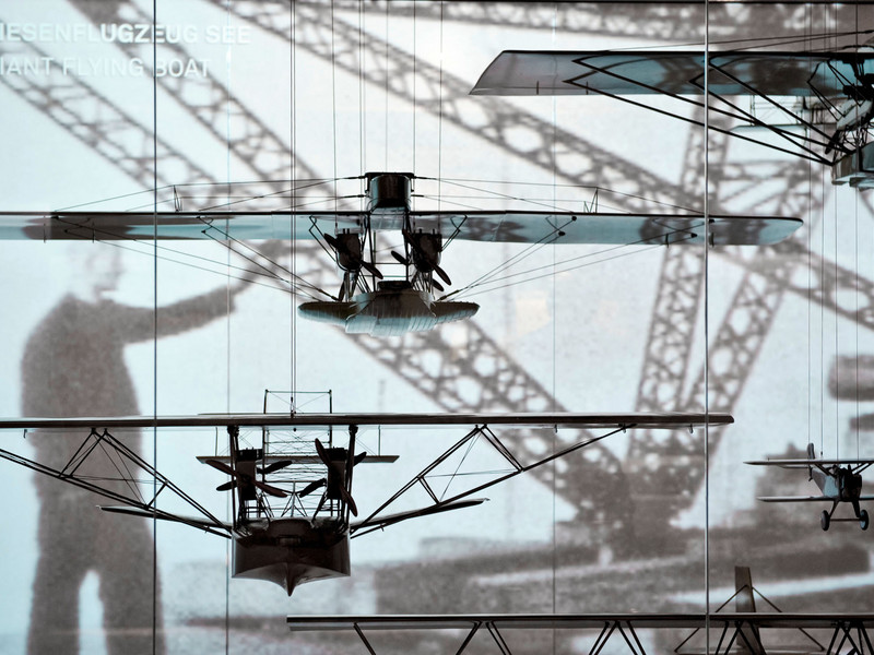 Mehrere Modellflugzeuge hängen vor einer Glaswand mit Fotomotiv von Baukränen und der Silhouette einer Person – Teil einer Ausstellung im Dornier Museum