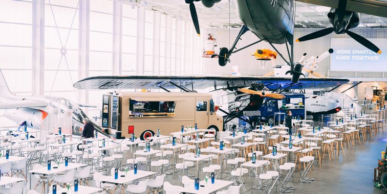Großer, heller Veranstaltungsbereich mit vielen weiß gedeckten Tischen und Stühlen zwischen historischen Flugzeugen in einer modernen Halle mit Glasfassade.