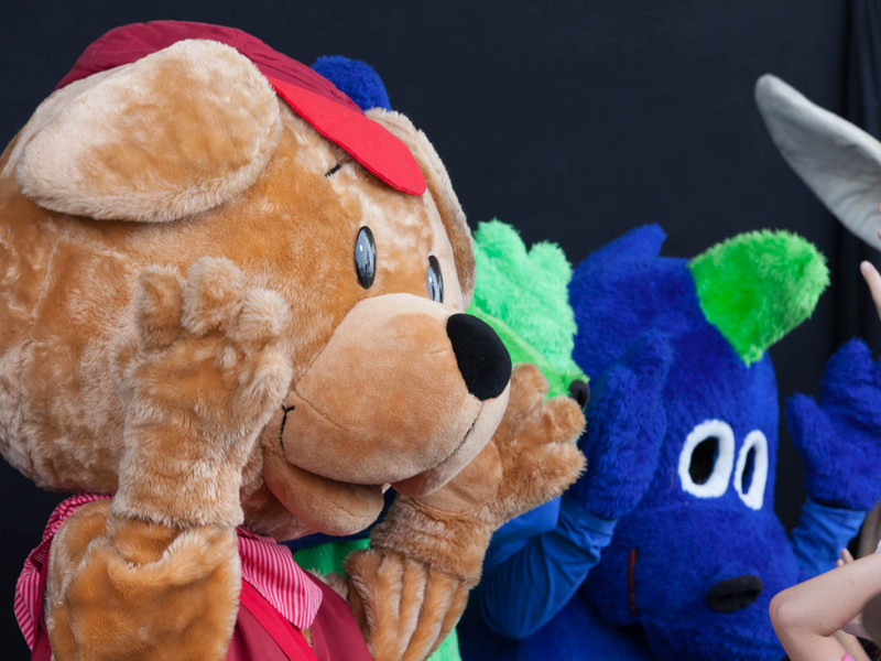 Zwei Personen in bunten Tiermaskottchen-Kostümen, ein Bär und ein blaues Wesen mit großen Augen, bei einer Veranstaltung im Freien.