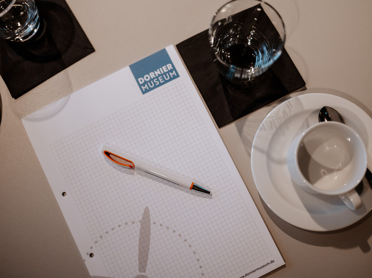 Nahaufnahme eines Konferenztisches mit Schreibblock, Stift, leerer Kaffeetasse und zwei Gläsern Wasser auf Untersetzern.