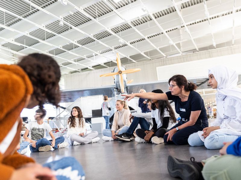 Schüler sitzen in einem Kreis auf dem Boden im Hangar.
