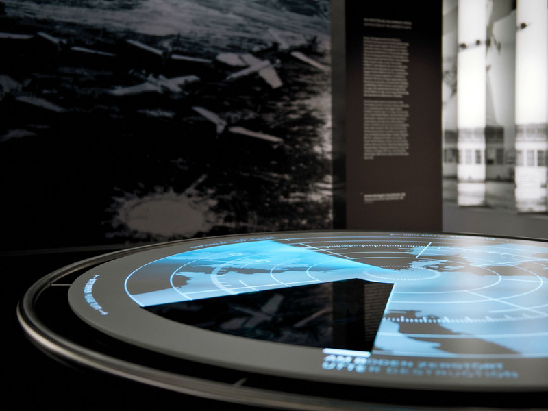 Runder, beleuchteter Tisch mit interaktiver Weltkarte vor dunklem Ausstellungshintergrund mit Flugzeuggrafiken und Lichtprojektionen im Dornier Museum