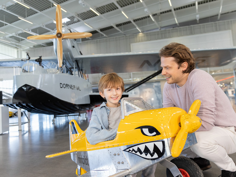 Ein Kind sitzt in einem Spielzeugflugzeug neben seinem Vater im Hangar mit Flugzeugen im Hintergrund.