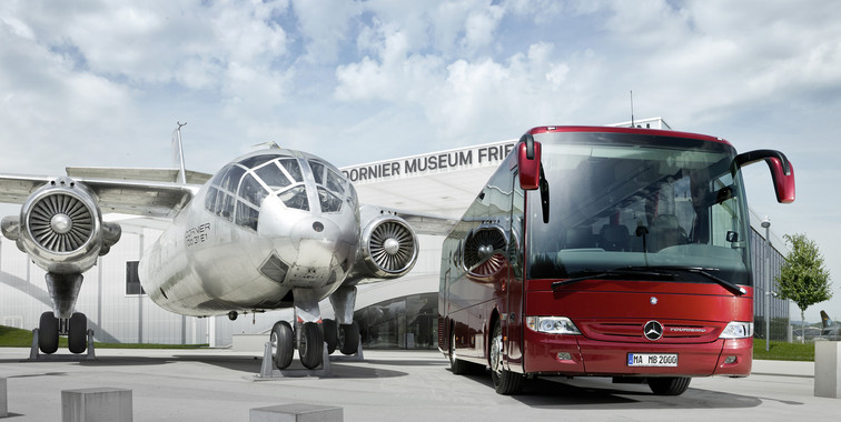 A silver plane in front of the Dornier Museum with a big red coach on the right of the picture.