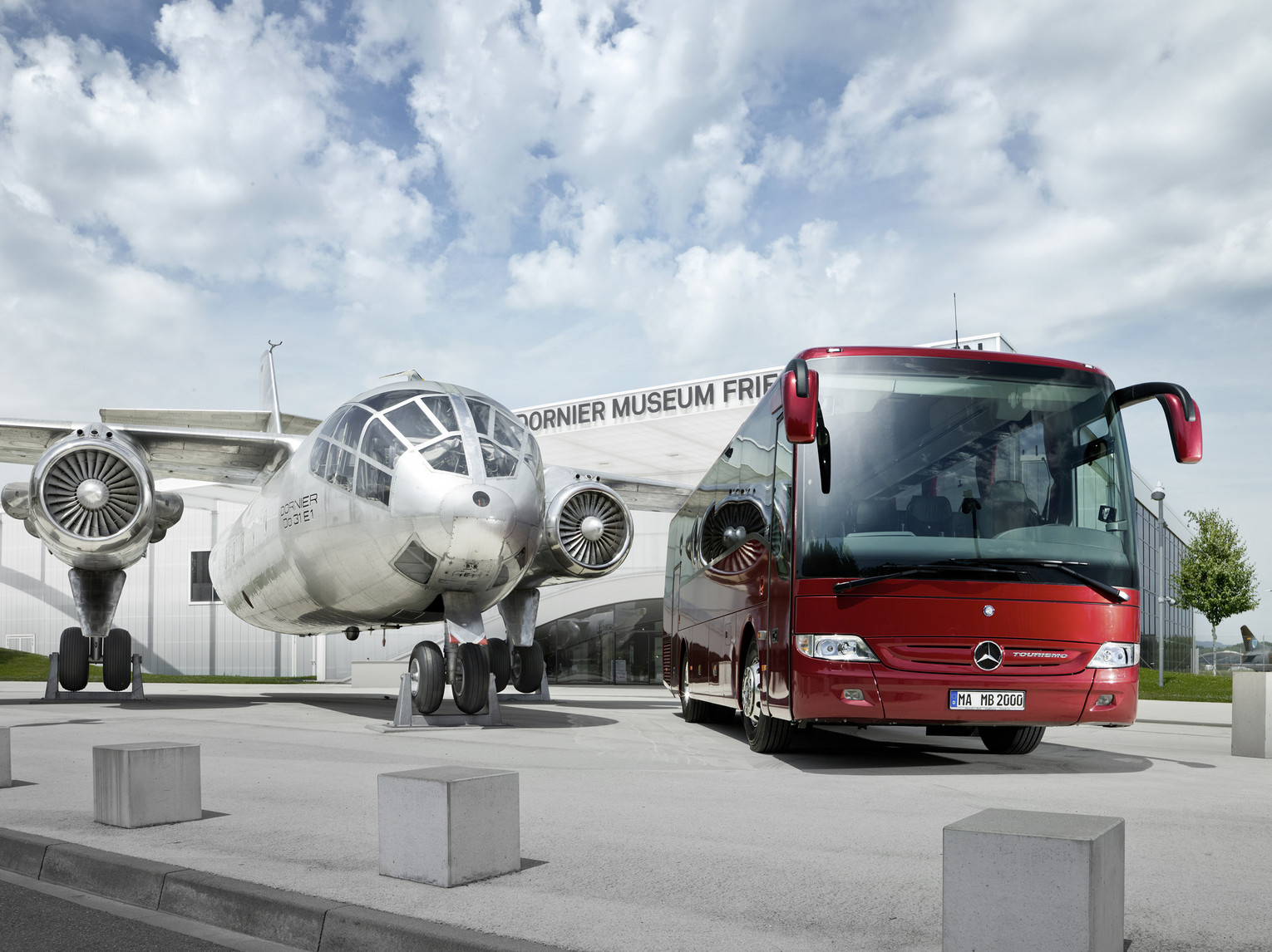 A silver plane in front of the Dornier Museum with a big red coach on the right of the picture.