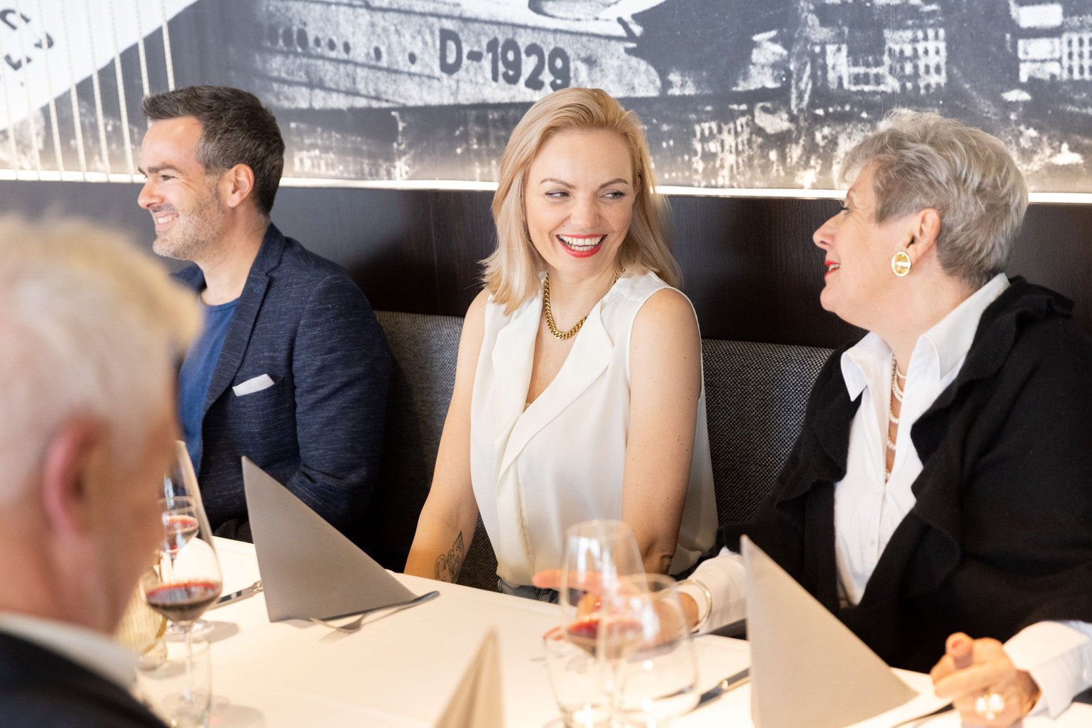Gruppe elegant gekleideter Menschen sitzt fröhlich an einem festlich gedeckten Tisch in einem Restaurant, im Hintergrund Wandbild mit historischem Flugzeug.