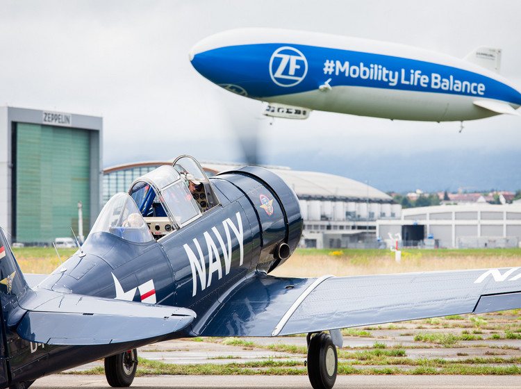Ein kleines Flugzeug steht mit laufendem Propeller auf der Startbahn, bereit zum Abflug. Im Hintergrund schwebt ein Zeppelin am Himmel.