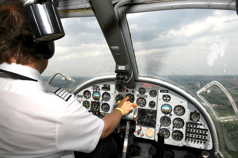 Pilot im Cockpit eines Flugzeugs mit Blick auf die Instrumente und nach vorne auf die Landschaft unter bewölktem Himmel.